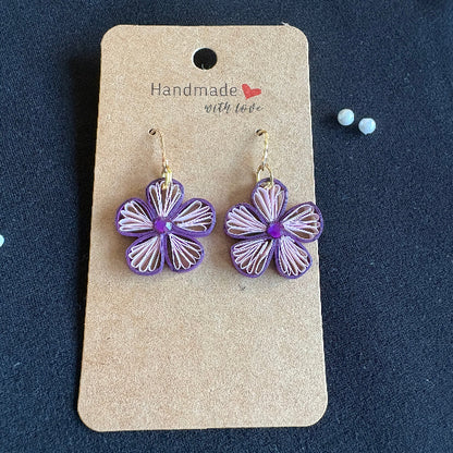 Purple flower-shaped earrings on a cardboard backing with 'Handmade with Love' tag against a dark background.