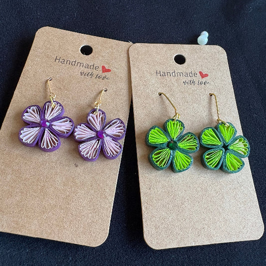 Pair of green and purple floral earrings on 'Handmade with Love' cards against a dark background.